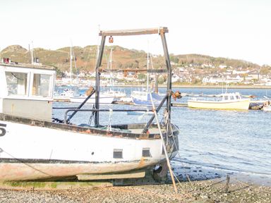 A white fishing boat on a rocky shore with boats and houses across the water at Dolwen in Penmachno