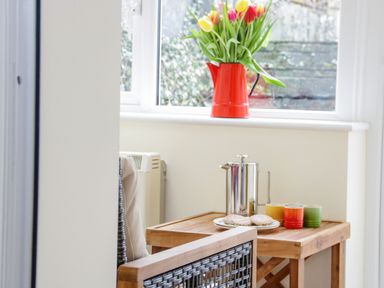 A wooden table with a metal coffee pot three colorful cups and cookies next to a window with a red vase of tulips and a wicker chair at Dolwen Penmachno