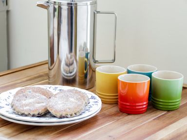 A wooden table with a silver French press four colorful mugs and a plate of pancakes dusted with powdered sugar