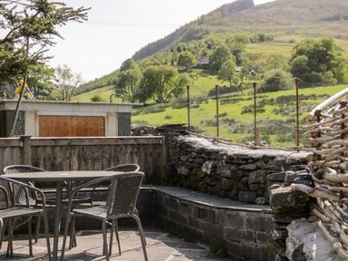 An outdoor patio with metal table and chairs surrounded by stone and wood fences with a hillside in the background at Dolwen Penmachno