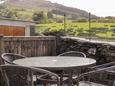 A glass table with four chairs on a stone patio with a wooden fence and green hills in the background at Dolwen in Penmachno