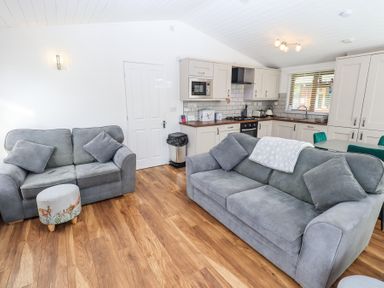 A living room with two grey sofas and a small ottoman in front of a kitchen with cabinets and a dining table at Grizedale Lodge in Langdale 18 White Cross Bay near Windermere