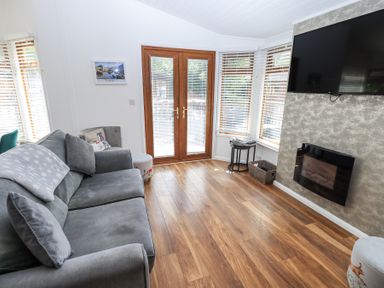 A living room with a gray sofa wooden floor a wall-mounted TV a fireplace and glass doors at Grizedale Lodge Langdale 18 White Cross Bay near Windermere