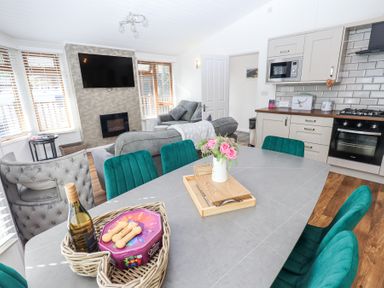 A living room and kitchen area with a dining table, green chairs, a TV mounted above a fireplace, and kitchen cabinets at Grizedale Lodge Langdale 18, White Cross Bay near Windermere