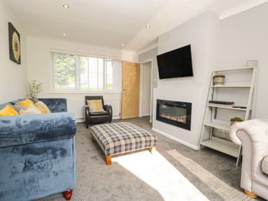 A living room with a blue sofa yellow cushions a plaid ottoman a wall-mounted TV an electric fireplace and a white shelving unit at 26 Hycemoor Way Bootle Near Millom