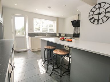 A kitchen with a central island and two stools next to a wall clock at 26 Hycemoor Way in Bootle Near Millom