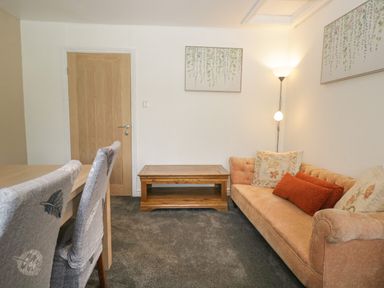 A living room with a wooden coffee table a beige sofa with cushions and two framed artworks on white walls at 26 Hycemoor Way in Bootle Near Millom
