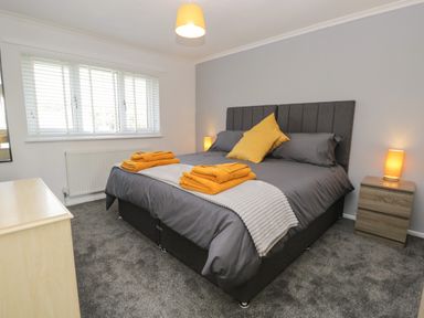 A bedroom with a double bed gray bedding yellow towels and pillows bedside table with lamp and a window at 26 Hycemoor Way Bootle Near Millom