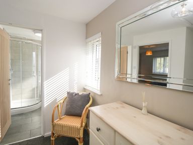 A small room with a wicker chair with a cushion a dresser with a mirror and an open door showing a bathroom with a shower at 26 Hycemoor Way in Bootle Near Millom