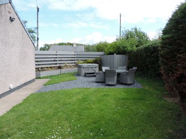 A backyard with a grass lawn a patio set with six chairs and a table a raised flower bed and a gray fence at 26 Hycemoor Way Bootle Near Millom