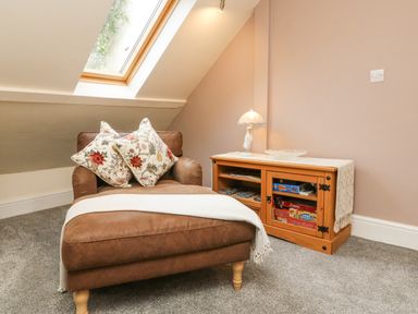 A brown armchair with two floral cushions and a white throw beside a wooden cabinet with board games and a lamp at Nattaden Fourwinds Meldon near Okehampton