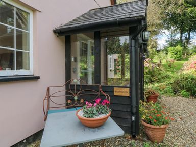 A small porch with a bench and a table holding a flower pot outside a house with window and garden at Nattaden Fourwinds Meldon near Okehampton