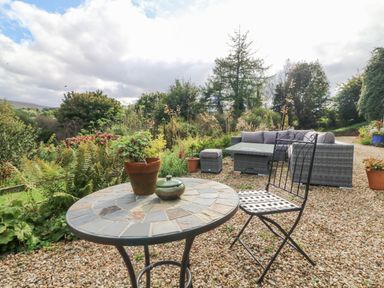 An outdoor patio with a round tiled table with plant pots a metal chair and a wicker sofa set with cushions among greenery at Nattaden @ Fourwinds in Meldon near Okehampton