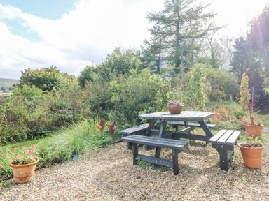 An outdoor seating area with a wooden picnic table and benches on gravel surrounded by potted plants and greenery at Nattaden @ Fourwinds in Meldon near Okehampton