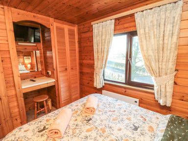 A bedroom with a bed with animal print bedding two rolled towels on it a wooden vanity with a stool a TV mounted above the vanity and a window with curtains at Holly Lodge in Liverton