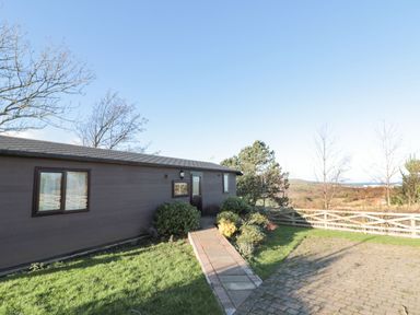 A brown building with windows and a door surrounded by grass bushes a paved path and a wooden fence with trees and hills in the background at Holly Lodge in Liverton