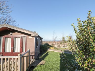 A wooden lodge with glass doors and a fenced deck surrounded by grass and bushes at Holly Lodge in Liverton
