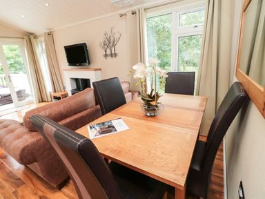 A dining area with a wooden table and black chairs next to a living room with brown sofas and a wall-mounted TV at Number 43 Burnside Lodge in Felton