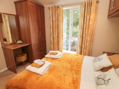 A bedroom with a bed covered in an orange blanket and pillows next to a window with orange curtains and a wooden wardrobe with a dressing table at Number 43 Burnside Lodge in Felton