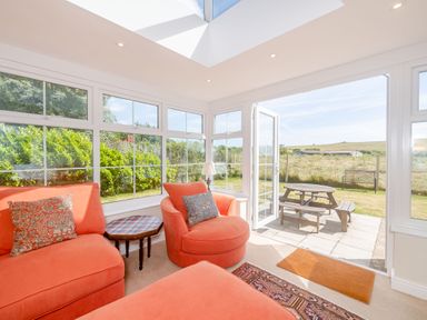 A sunroom with coral armchairs a patterned side table and glass doors opening to a garden with picnic bench at West Bay Holiday Home in West Bay