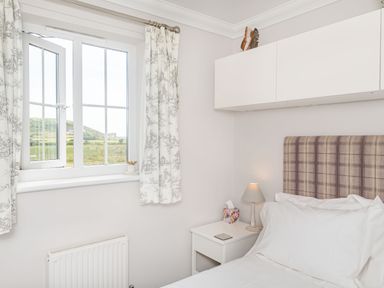 A bedroom with a window with patterned curtains a bedside table with a lamp a tissue box and a bed with a plaid headboard at West Bay Holiday Home in West Bay