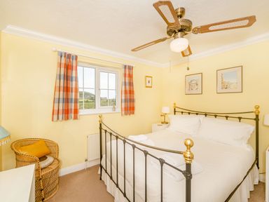 A bedroom with a metal bed frame white bedding bedside tables lamps a ceiling fan and a window with checkered curtains at West Bay Holiday Home in West Bay