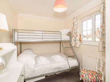 A bedroom with a metal bunk bed white bedding a chair with a colorful cushion and a window with patterned curtains at West Bay Holiday Home in West Bay