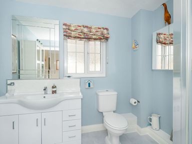 A bathroom with a white sink and cabinet a toilet a window with patterned curtains and a mirrored cabinet at West Bay Holiday Home in West Bay