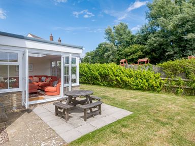 A garden with a picnic table on a patio and an open door leading to a living room with a red sofa at West Bay Holiday Home in West Bay
