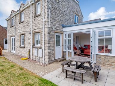 Stone house with open white framed doors leading to a room with red couches and wooden picnic table outside on a patio at West Bay Holiday Home in West Bay