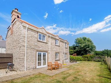 A stone exterior of a house with two wooden chairs on a patio and a lawn with trees at West Bay Holiday Home in West Bay