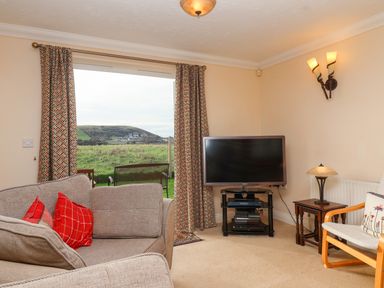 A living room with a sofa, television on a stand, armchair, side table, lamp, and large window with curtains at West Bay Holiday Home in West Bay