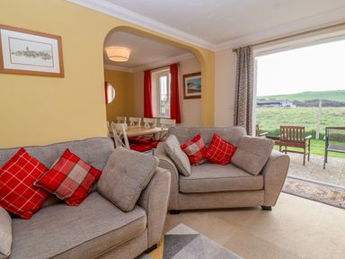 A living room with gray sofas and red cushions next to a dining area with white chairs and a window at West Bay Holiday Home in West Bay