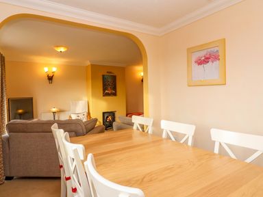 A dining area with a wooden table and white chairs leading to a living room with sofas a TV and a fireplace at West Bay Holiday Home in West Bay