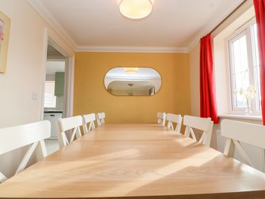 A dining room with a wooden table and white chairs near a window with red curtains at West Bay Holiday Home in West Bay