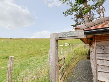 A wooden entrance gate with a sign that says Welcome to Y Caban next to a wooden building and a fenced grass field with sheep at Y Caban in Pontfadog near Llangollen