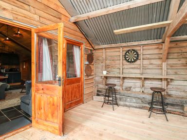 A rustic wooden room with a dartboard a wooden bar with two stools and an open door leading to a living room at Y Caban Pontfadog near Llangollen