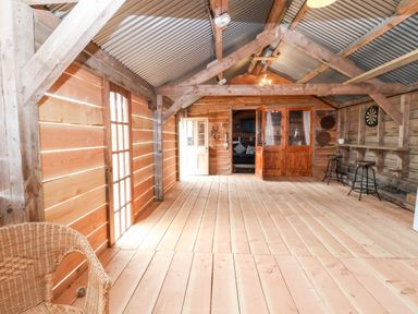 An empty wooden room with a wicker chair a dartboard two bar stools and double wooden doors at Y Caban Pontfadog near Llangollen
