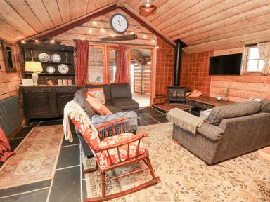 A living room with wooden walls and ceiling two gray sofas a rocking chair a wood stove and a black cabinet with plates at Y Caban Pontfadog near Llangollen