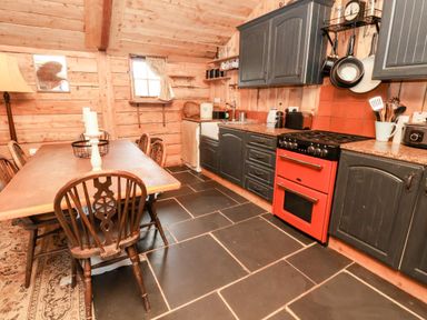 A kitchen with wooden walls and ceiling a red stove black cabinets a wooden dining table with chairs and a window at Y Caban in Pontfadog near Llangollen