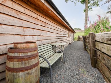 A wooden bench and table beside a wooden barrel on gravel next to a wooden building and flower bed at Y Caban in Pontfadog near Llangollen