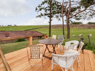 A wooden deck with a round table and mismatched chairs overlooking a green field with sheep at Y Caban in Pontfadog near Llangollen