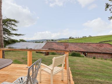 A wooden deck with a round table and chairs overlooking a grassy hill and buildings with tiled and metal roofs at Y Caban in Pontfadog near Llangollen