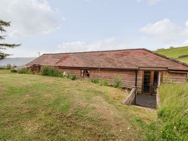 A wooden single-story building with a sloped roof surrounded by grass and plants at Y Caban in Pontfadog near Llangollen