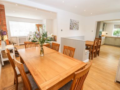A dining room with a wooden table and chairs a floral arrangement and a view into a kitchen area at 17 Sands Close in Broadway
