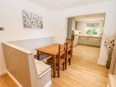 A dining area with wooden table and chairs next to a built-in bench with cushions and an open view of the kitchen at 17 Sands Close in Broadway
