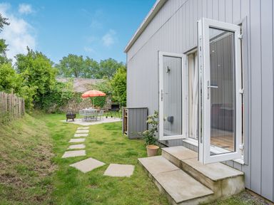 A garden with stepping stones leading to a patio with table chairs and umbrella beside a building with open glass doors at Caldey Lodge Bier Wood Lodges in Manorbier