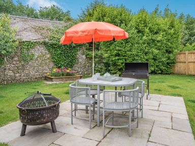 An outdoor patio area with a table and chairs under an orange umbrella a fire pit and a barbecue grill in a garden at Caldey Lodge (Bier Wood Lodges) in Manorbier