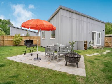 A backyard with a patio table and chairs an orange umbrella a barbecue grill and a fire pit at Caldey Lodge (Bier Wood Lodges) in Manorbier