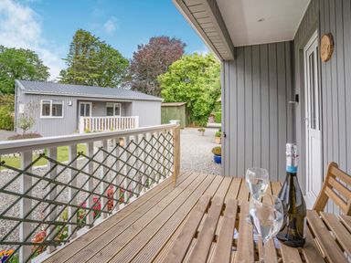 A wooden porch with a table holding a bottle and two glasses outside a gray cabin at Caldey Lodge (Bier Wood Lodges) in Manorbier